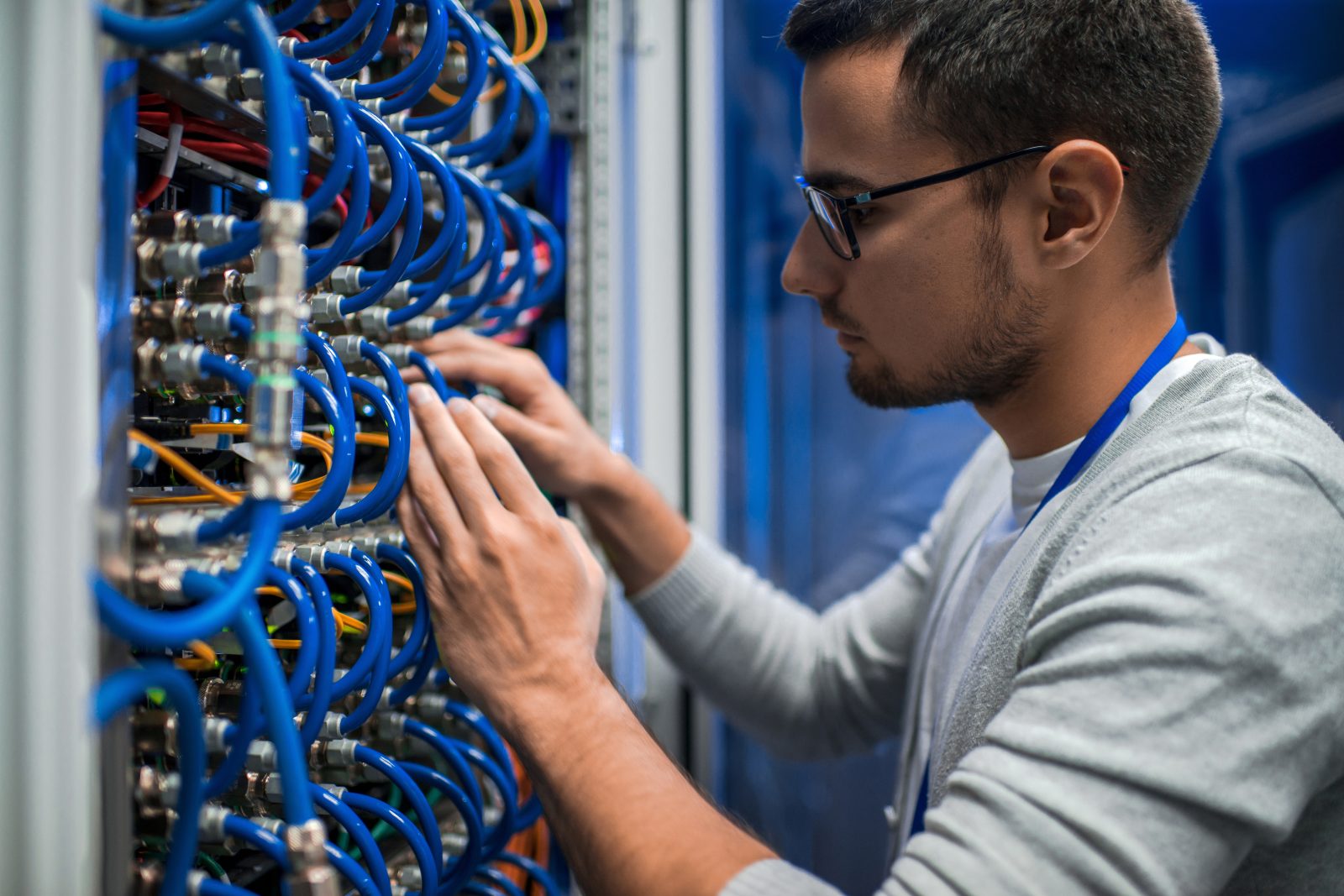 A man in a white t-shirt, gray sweater, blue lanyard, and glasses adjusts the blue cables of a server rack