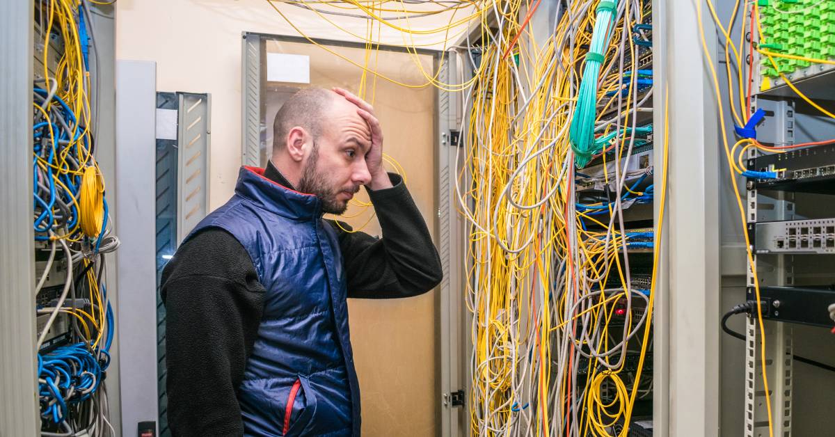 A man holds his hand to his head and looks overwhelmed by the state of a data room, which has poor cable management.