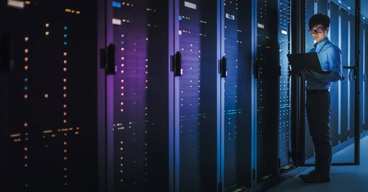 A man in a blue button-down shirt holds a laptop next to a row of server racks in a dark data center.