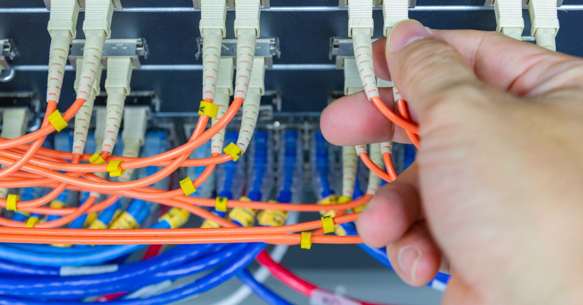 A hand plugs an orange fiber cable into a switch port. There are other orange, blue, and red cables around it.