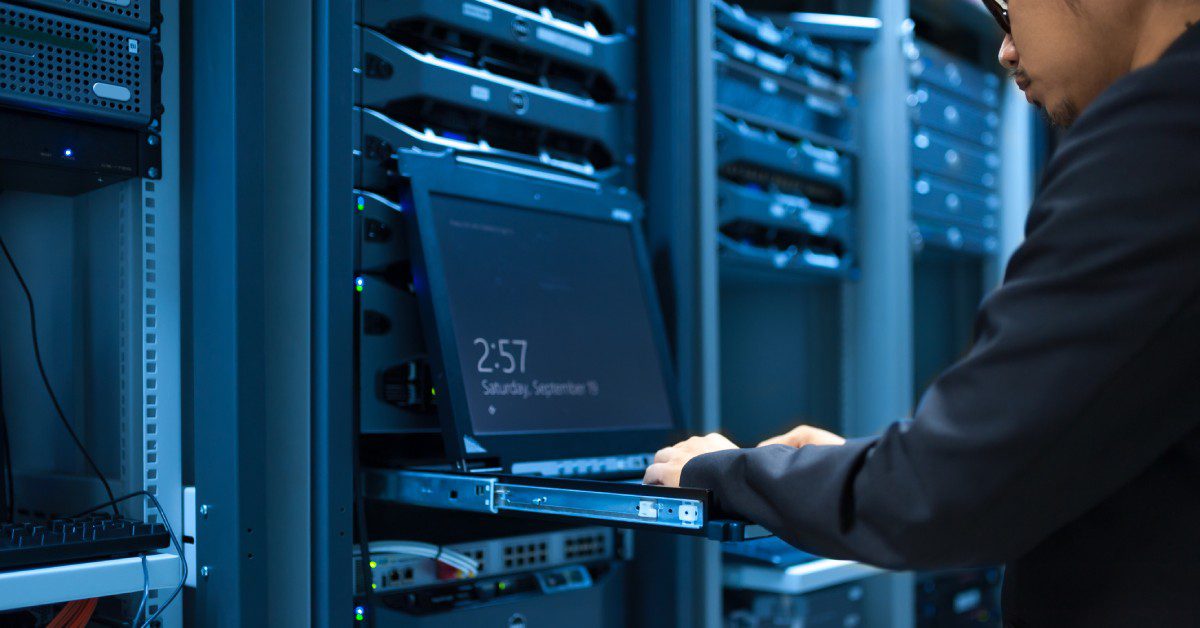 A man types on a laptop open to a screensaver resting on a pulled-out shelf in the aisle of a data center.