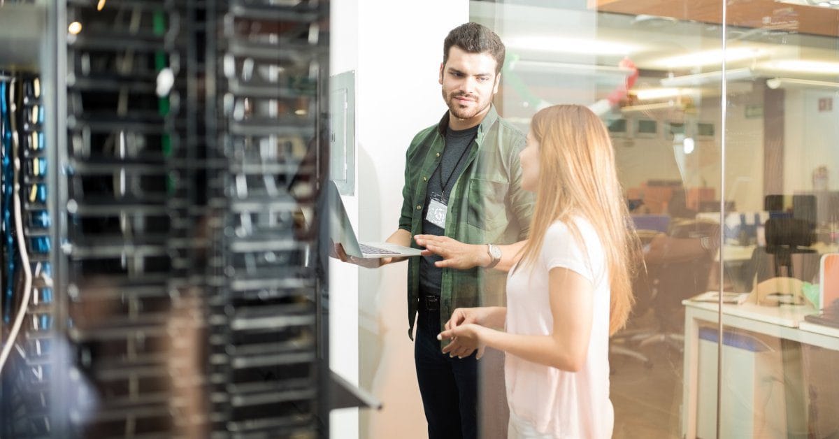 A woman in a white shirt speaking to a bearded man who is holding a laptop. They are in a server room next to an office area.