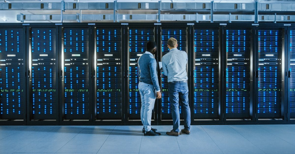 Two men in business casual attire standing in a data center with their backs to the camera. One is holding a laptop.