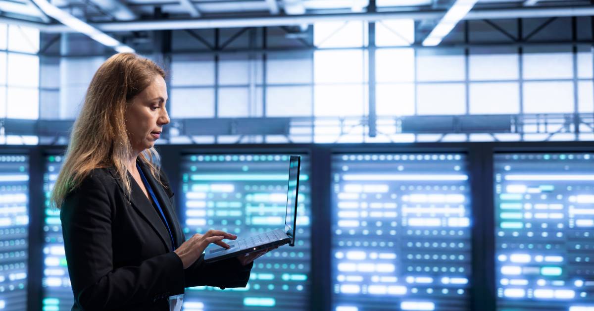 A woman stands holding a laptop and using the mousepad in front of servers emitting blue light in a data center.