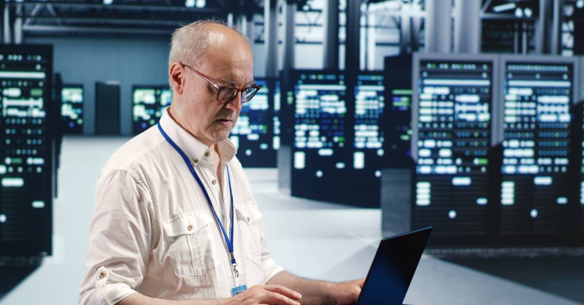 A man wearing eye glasses and a blue lanyard works on a laptop inside of a data center with servers behind him.