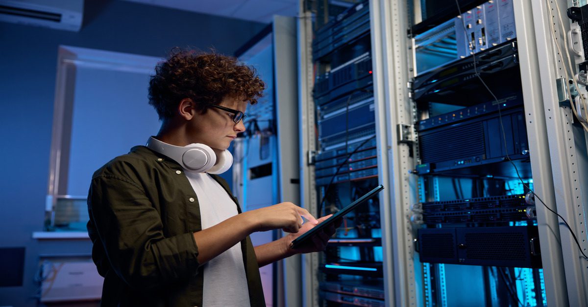 A man wearing headphones around his neck holds a tablet while standing in front of a data server rack.